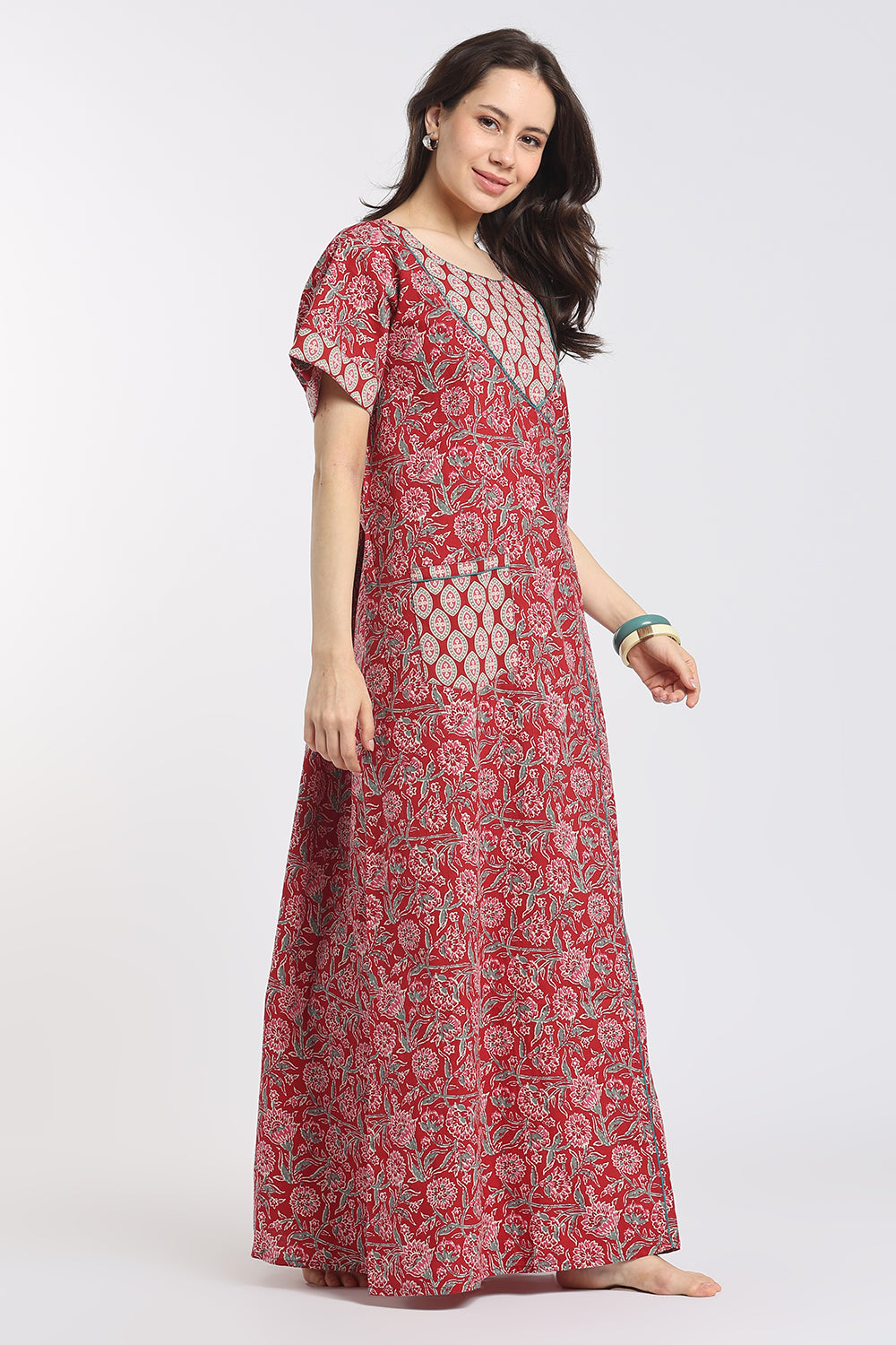 A woman modeling a red and pink floral block-printed maxi dress with short sleeves and a round neckline, set against a plain light gray background.