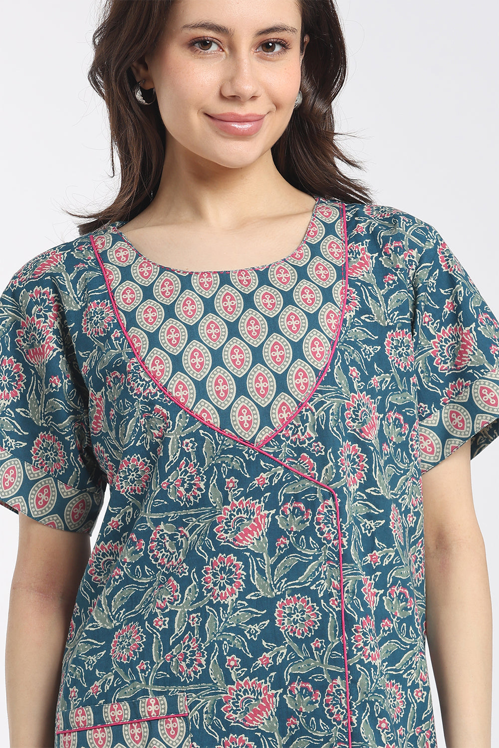 A woman wearing a short-sleeve printed tunic top with a teal blue base, pink and white floral motifs, and pink contrast stitching, shown against a plain light gray background.