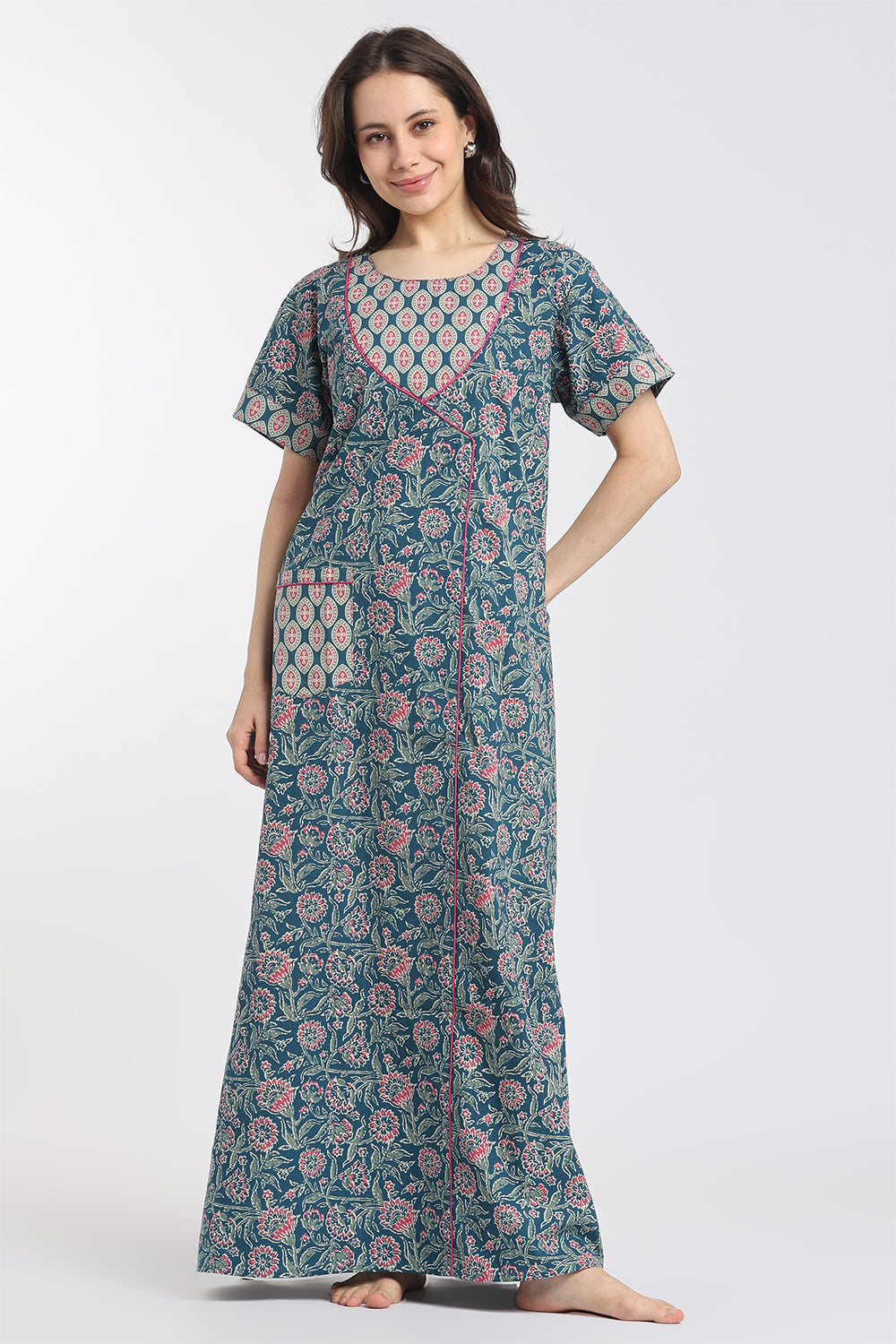A woman modeling a long-sleeved, floor-length nightgown with a teal blue base and pink and white floral block print, standing against a light gray studio background.