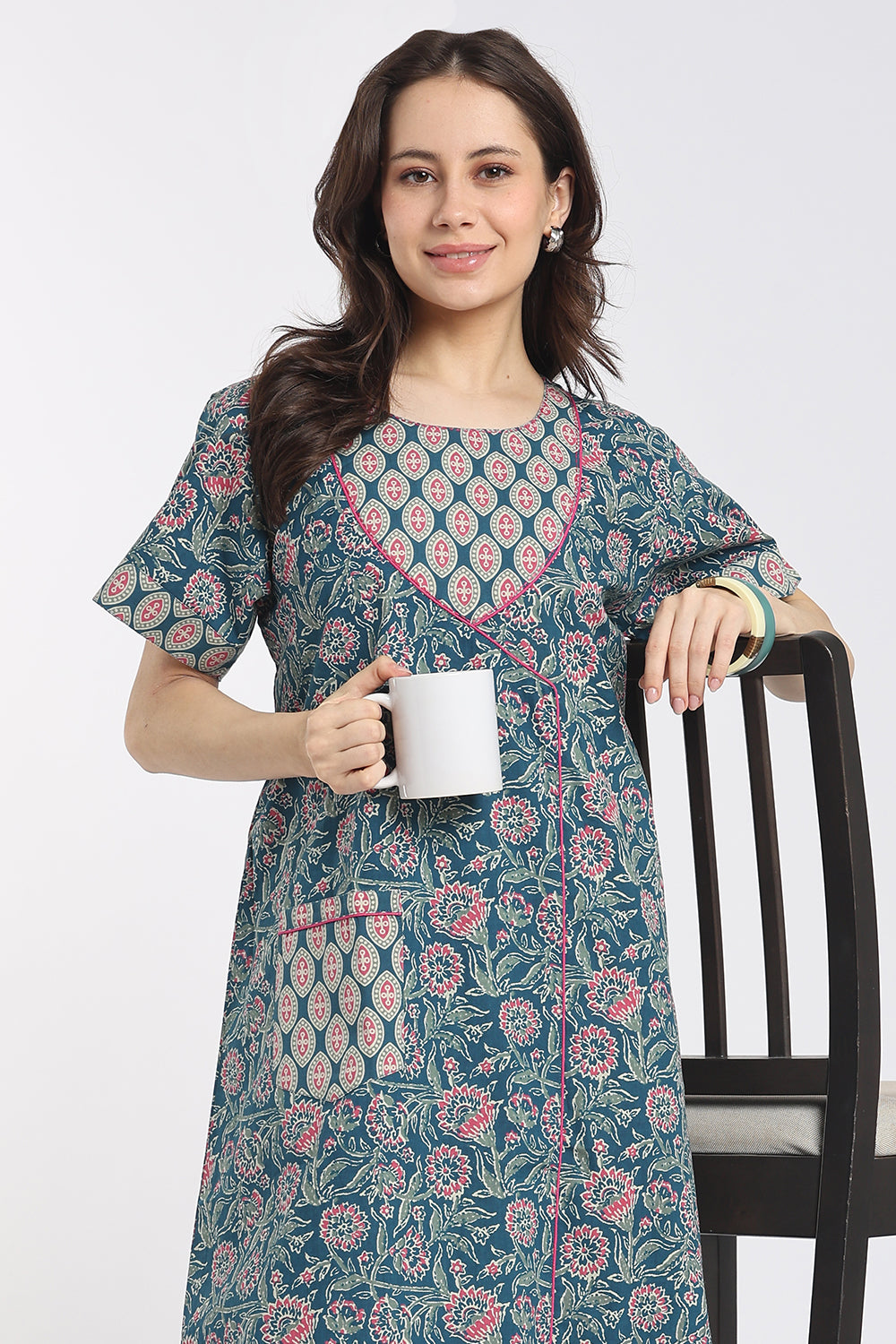 A woman wearing a short-sleeved teal blue printed kurta dress with pink and beige floral motifs, holding a white mug next to a black wooden chair against a light gray background.