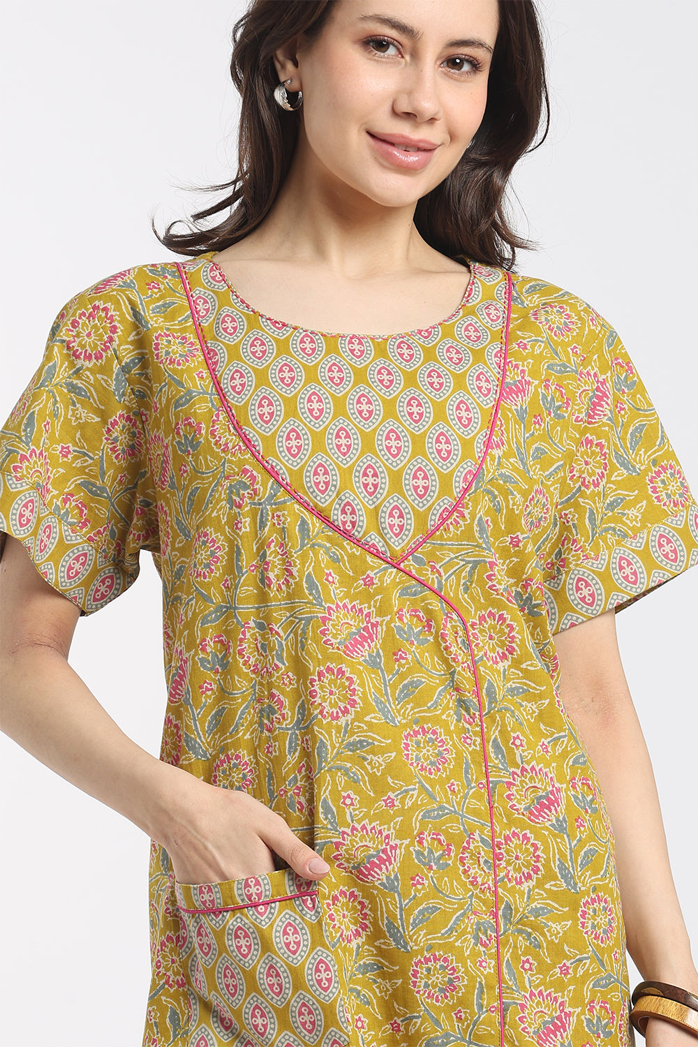 A woman wearing a mustard yellow short-sleeve tunic top with a pink and green floral print and pink contrast piping, shown against a light gray studio background.
