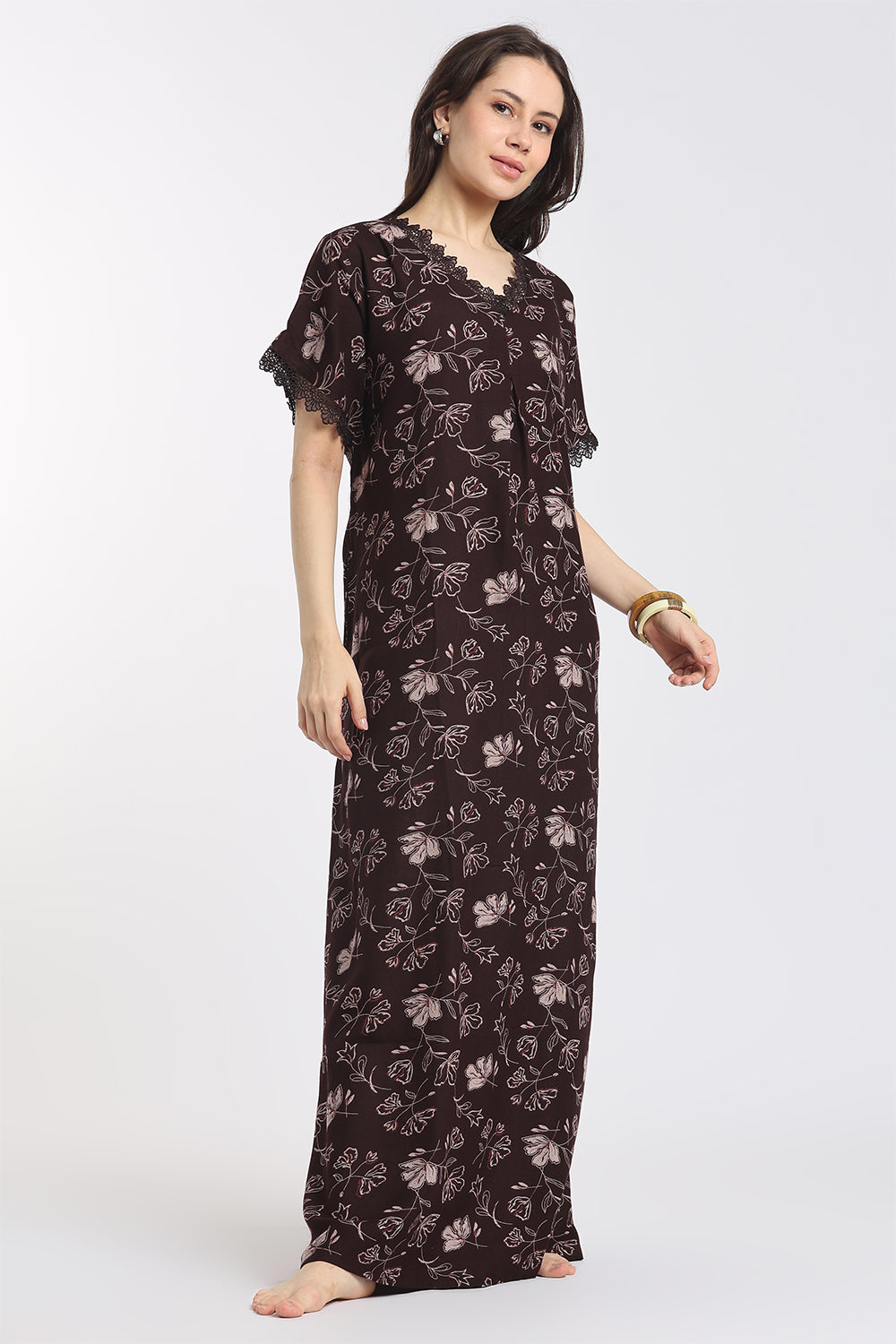 A woman modeling a long, dark brown nightgown with a light floral print and ruffled short sleeves, standing against a plain light gray background.
