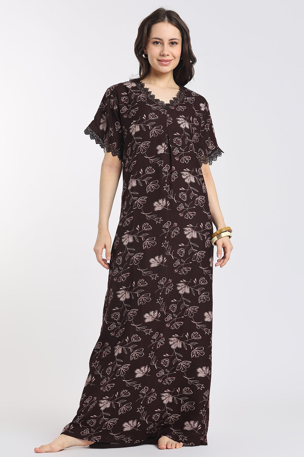 A woman modeling a long, loose-fitting nightgown or kaftan dress in dark brown with a light beige floral pattern, featuring short flutter sleeves with lace trim and a scalloped neckline.