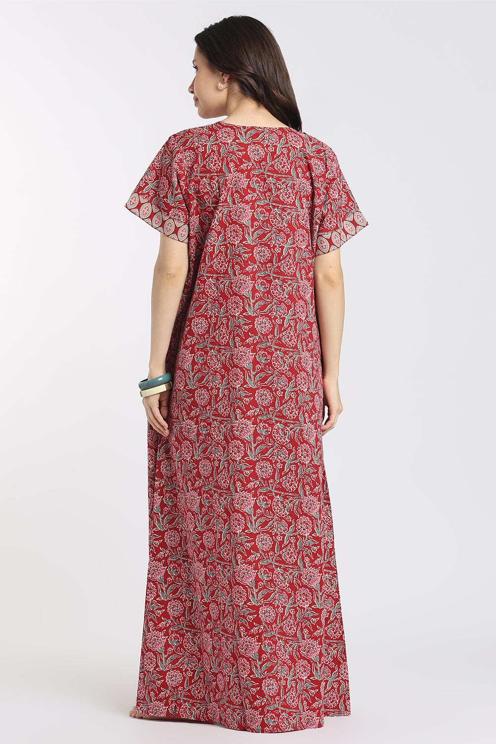 A woman viewed from the back wearing a long red dress with a white floral block print and short flutter sleeves, set against a plain light gray studio background.
