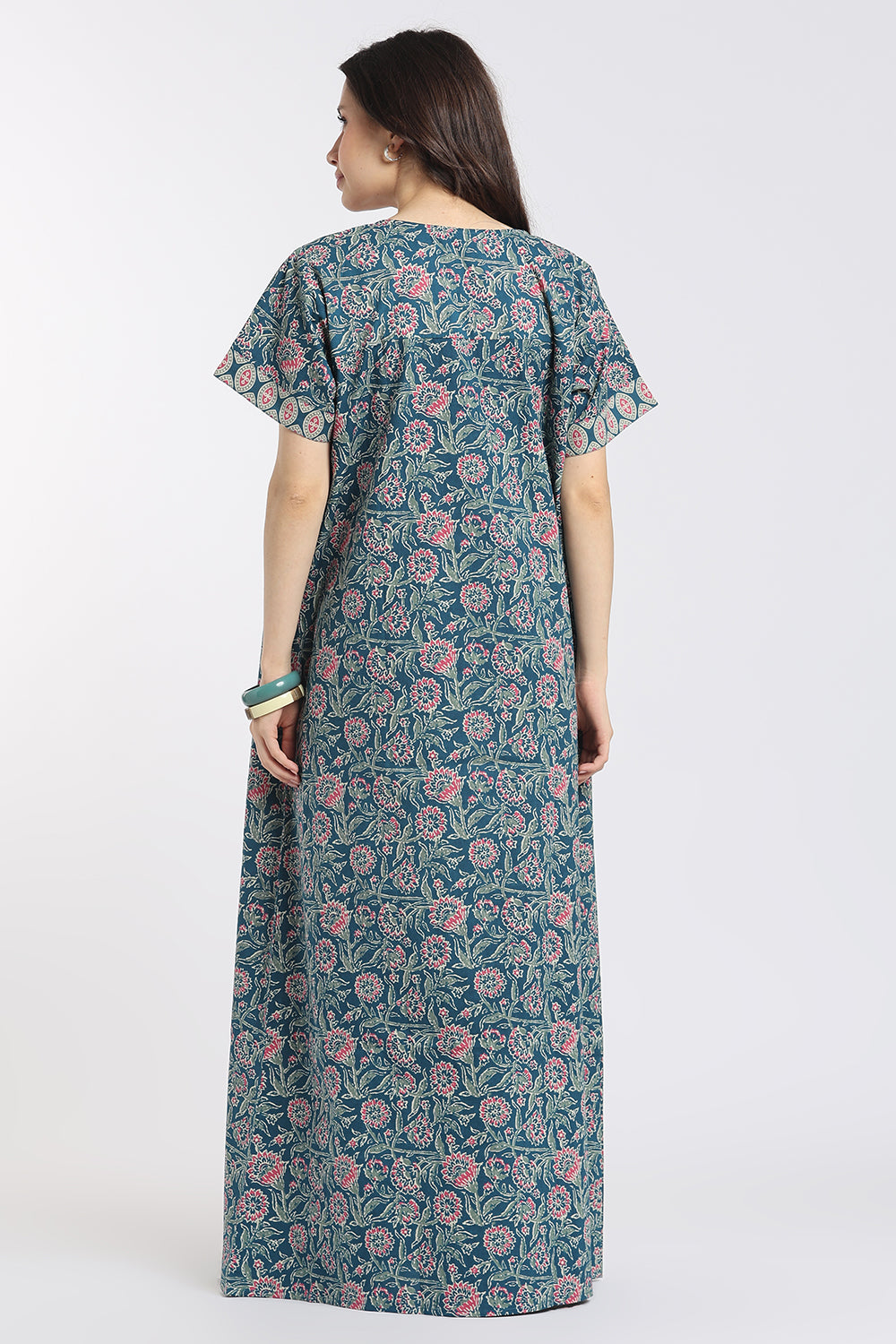 A woman viewed from the back wearing a long-sleeved maxi dress with a teal blue and pink floral block print pattern, set against a plain light gray studio background.