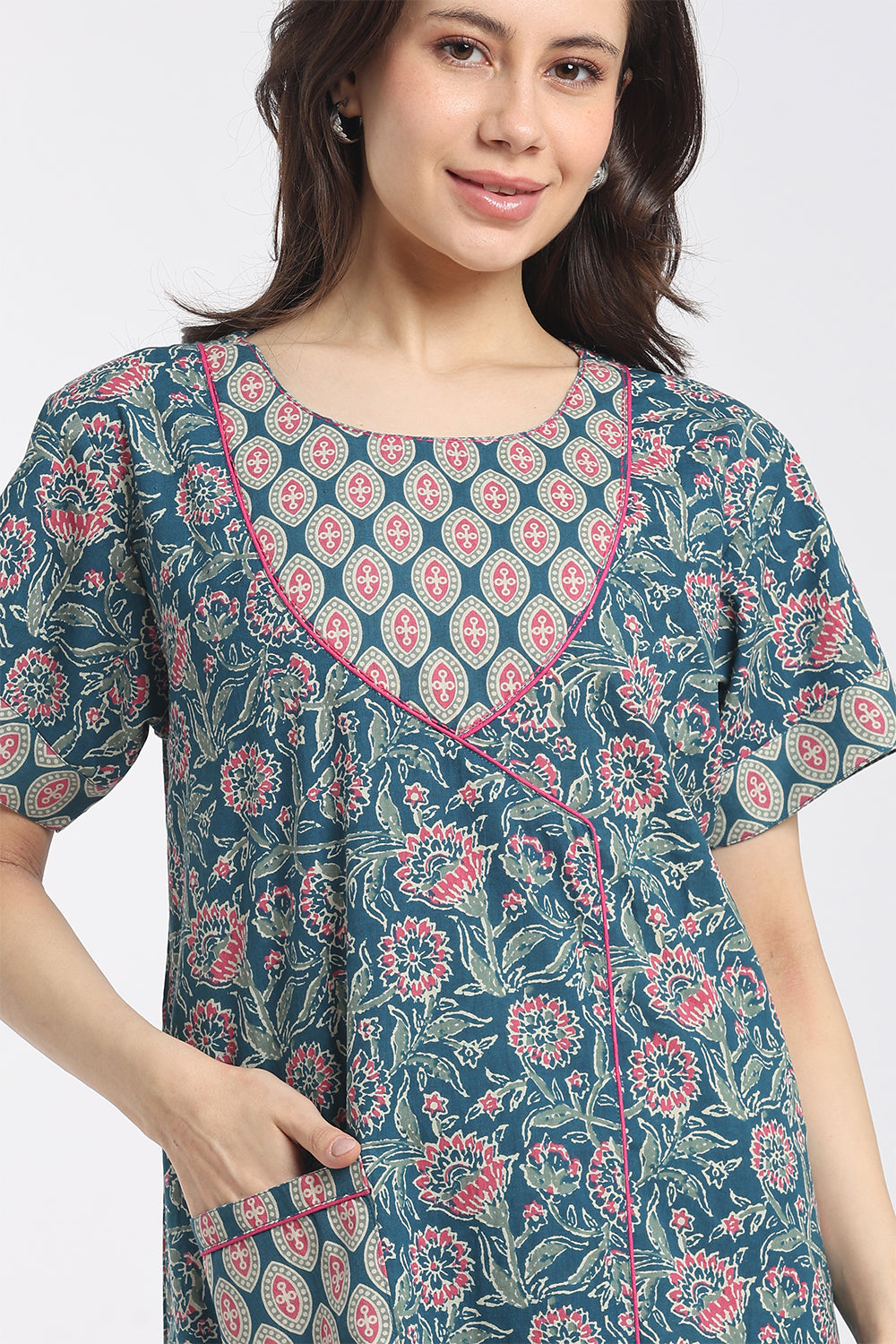 A woman wearing a short-sleeve wrap-style tunic dress with a blue-green base and pink and white floral block print, featuring a pink embroidered neckline.