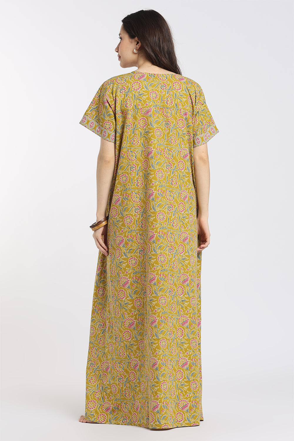 A woman viewed from the back wearing a loose, short-sleeve maxi dress with an all-over paisley print in olive green, pink, and yellow, set against a plain light gray studio background.