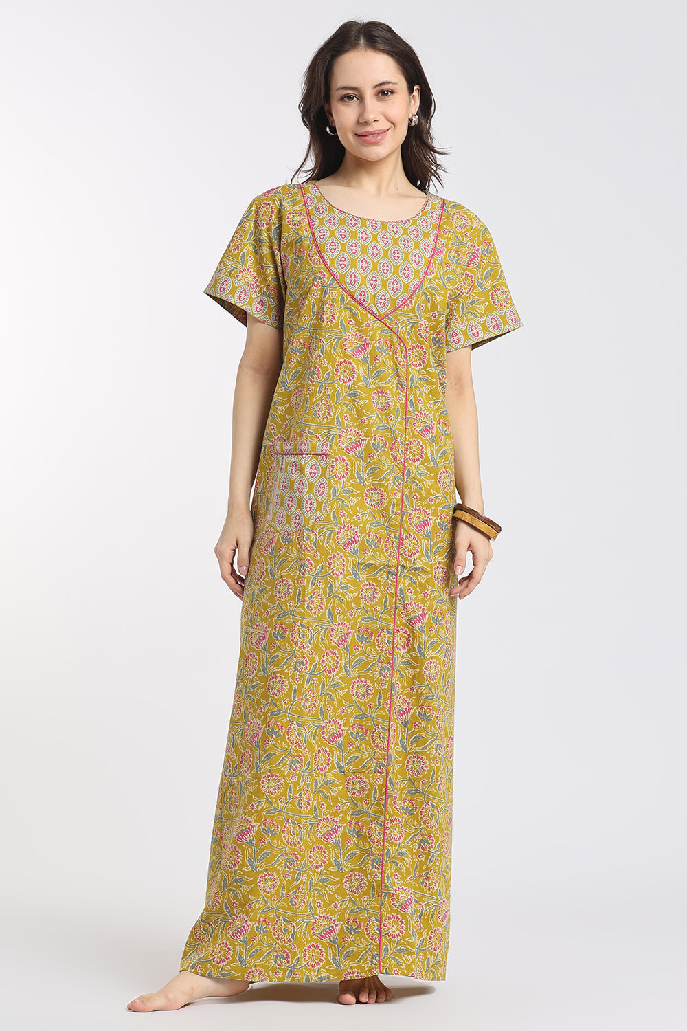 A woman modeling a long-sleeved, floor-length printed nightgown or kaftan in a yellow-green base with pink and orange floral patterns, set against a plain light gray studio background.