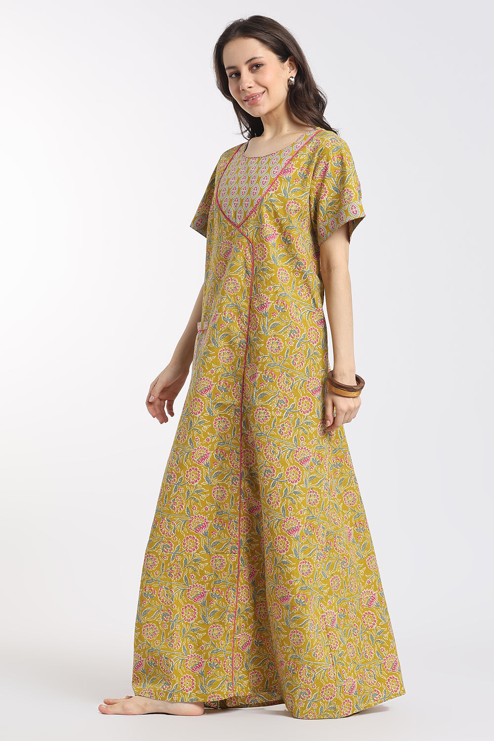 A woman modeling a long, loose-fitting yellow printed kaftan dress with pink and green floral patterns, standing against a plain white studio background.