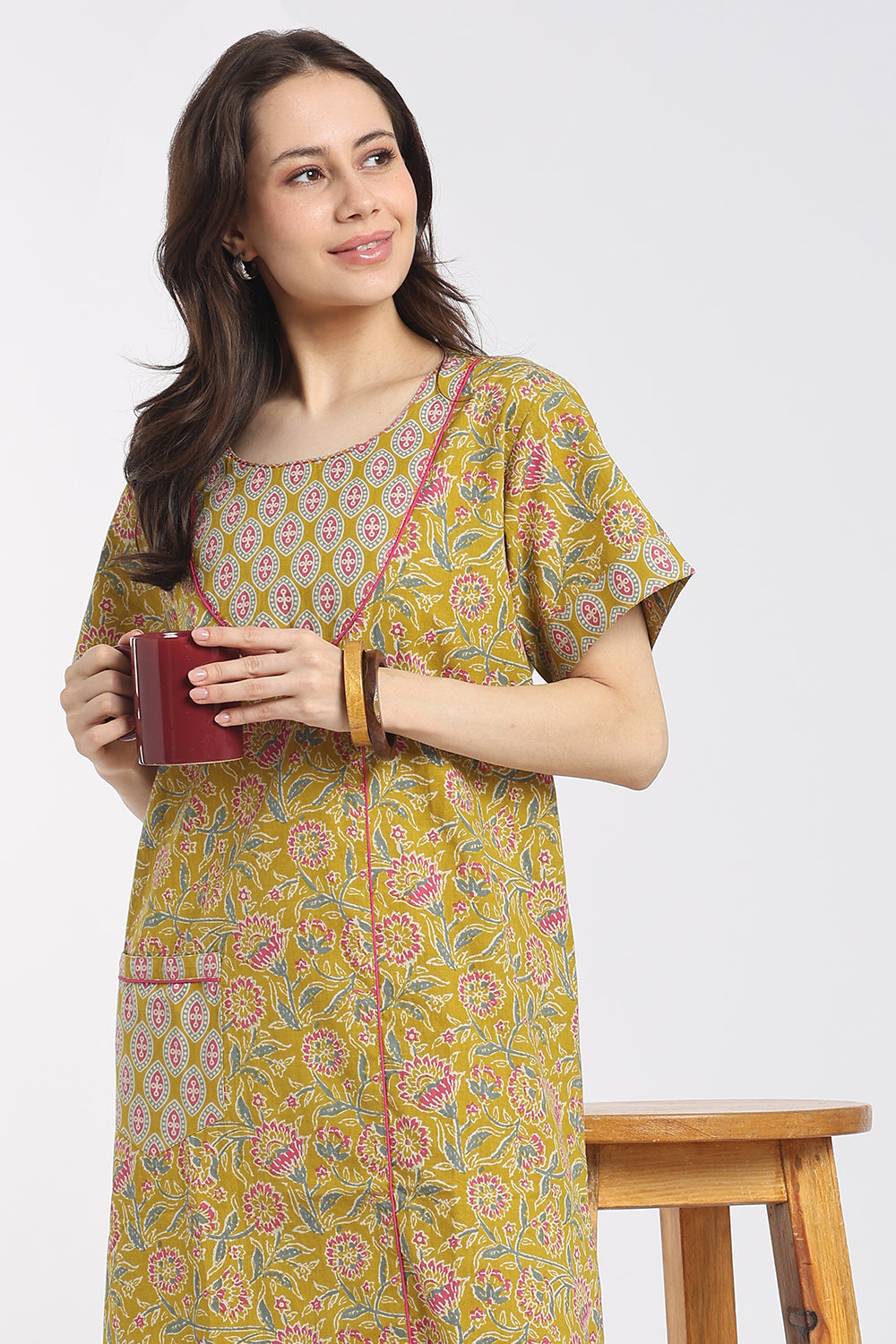 A woman wearing a yellow kurta with a multicolor floral print and holding a dark red mug, standing next to a wooden stool against a light gray background.