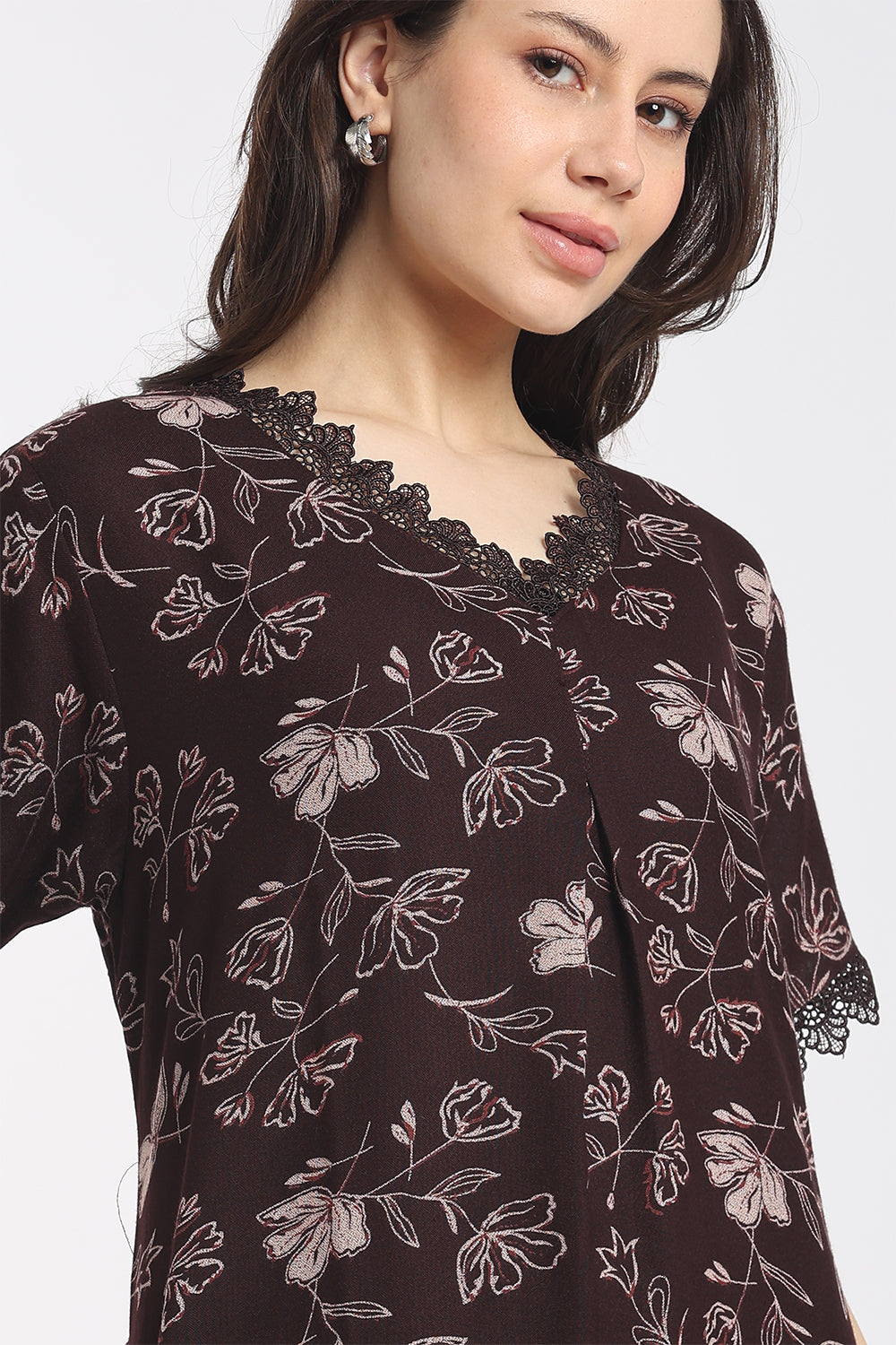 A close-up of a woman wearing a dark brown floral print top with black lace trim at the V-neck and sleeve cuffs, set against a plain light gray background.