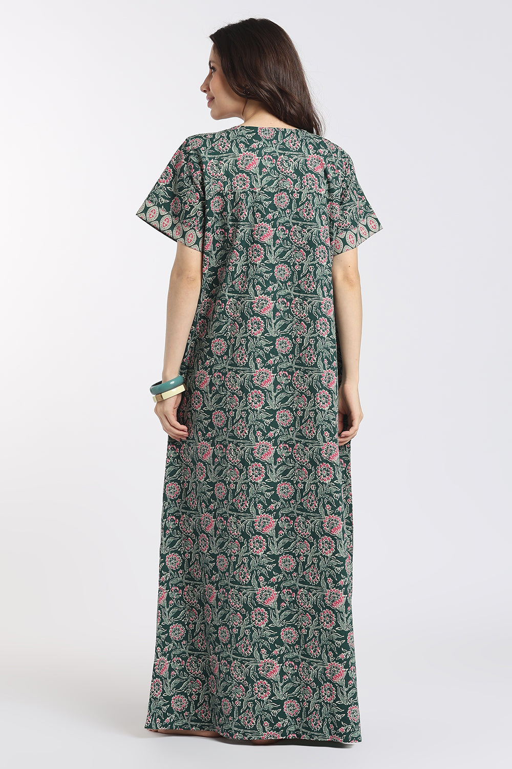 A woman viewed from the back wearing a long, short-sleeved dress with a green base and an all-over pink and white floral block print, set against a plain light gray studio background.