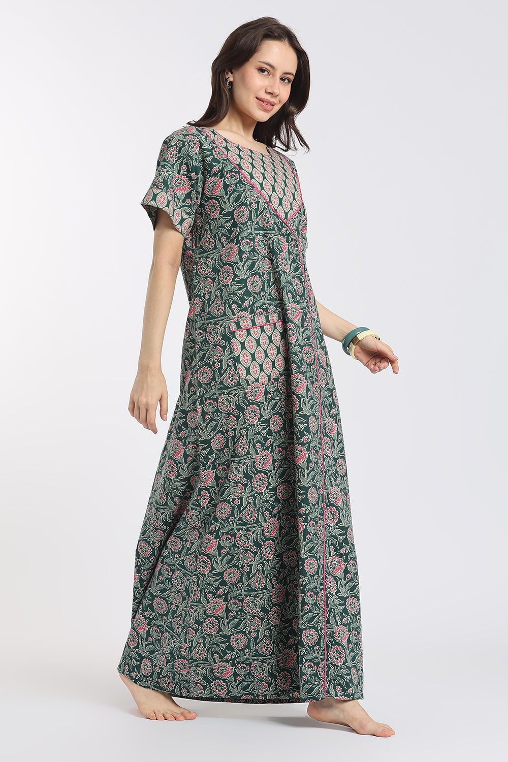 A woman modeling a long-sleeved, floor-length maxi dress with a green base and a pink floral block print pattern, set against a plain light gray studio background.